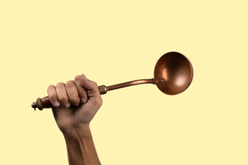 Black male hand holding a brass vintage kitchen ladle on yellow background
