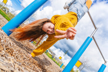 Cheerful girl hanging upside down on gymnastics bar and showing peace sign