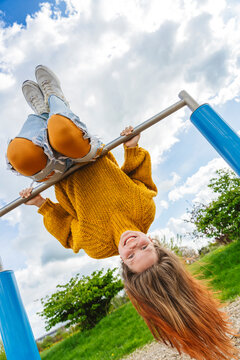 Playful girl hanging upside down on gymnastics bar under cloudy sky