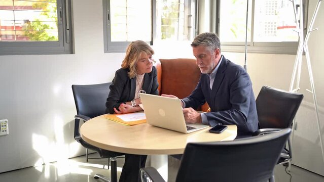 Two senior business professionals analyzing documents together at a round table with a laptop in a sunlit office environment. 