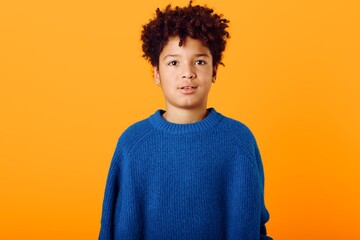 Cheerful young african american boy in vibrant blue sweater standing against a bright orange background with big smile