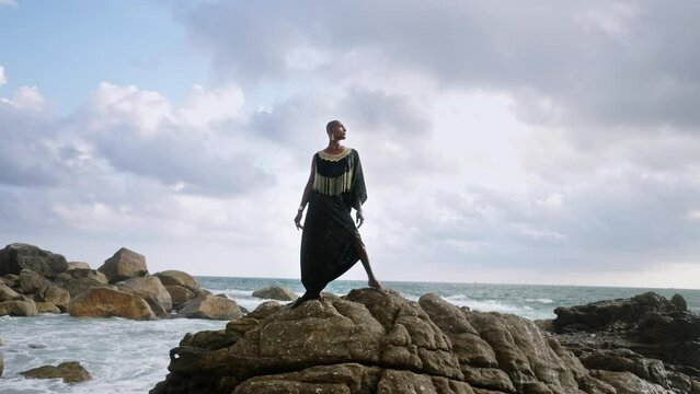 Godlike gender fluid person of color in luxury dress, jewelry stands on cliff peak at scenic ocean. Pride extravagant fashion model in epic outfit posing like goddess in tropics. Olymp concept.