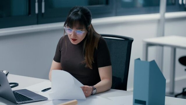 Close up of a young beautiful dark haired woman working at her papers with laptop in office, taking her glasses off, being exhausted, tired and stressed at work in slow motion