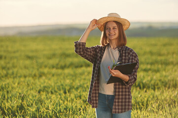 Front view, standing and holding documents. Young woman is on the beautiful agricultural field at daytime