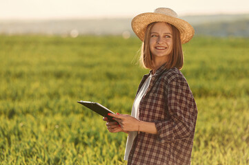 Positive mood, with straw hat and with documents. Young woman is on the beautiful agricultural field at daytime