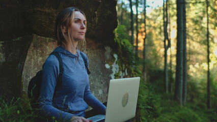 A young woman dressed in earthy tones is sitting cross-legged on mossy ground in a mystical forest, typing on a laptop with rays of sunlight filtering through the canopy above her.