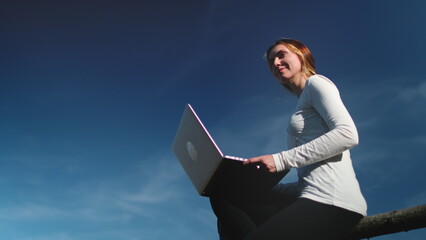 Young woman in modern clothing sits gracefully on a rustic wooden fence, absorbed in her laptop screen. The sun shines down on her as she navigates the digital world, blending nature and technology.