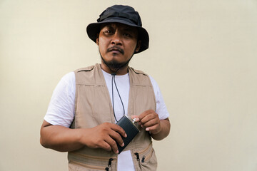 Macho asian man with beard and mustache wearing safari clothes isolated on beige background. Half body portrait of adult Southeast Asian man posing wearing vest, bucket hat and hip flask