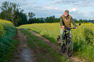Fototapeta premium An active senior cycles early in the morning on a sunny field path, surrounded by brightly blooming yellow rapeseed fields, enjoying the peace and quiet of the morning hours.