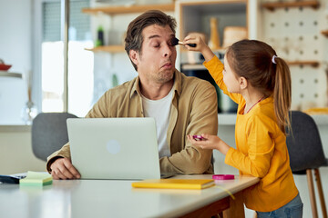 A working father letting his playful daughter put makeup on his eyes while he is working from home