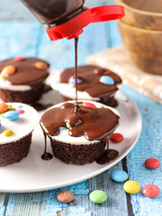Chocolate sauce is poured from a bottle onto a plate with cupcakes. The cupcakes have white icing and are topped with colorful candy-coated chocolates. In the background, a wooden bowl is partially.