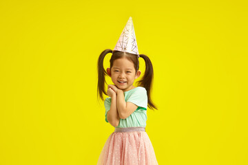 Asian little girl in party hat smiling coyly, cute placed palms of hands in front of herself face,...