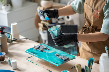 Female artist pours epoxy resin and smears it with stick on wooden board. Craftswoman using liquid...
