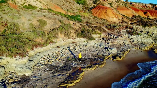 Aerial view of rugged limestone cliffs and coastal erosion at a beach with blue water and waves and a person in an orange jacket walking along the beach, Port Noarlunga, South Australia, Australia