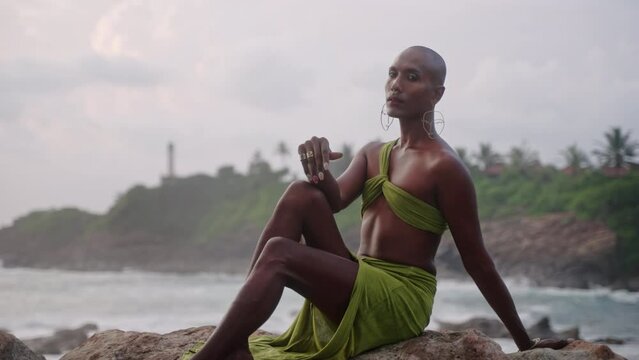 Gay bipoc man in luxury dress, boutique brass jewelry, makeup poses sitting above rocky ocean beach. Homosexual epatage fashion model in open outfit, nose-ring piercing, earrings, rings with gems.