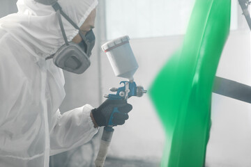 Car repair painter in protective overalls and painting a respirator part of car in the paint chamber