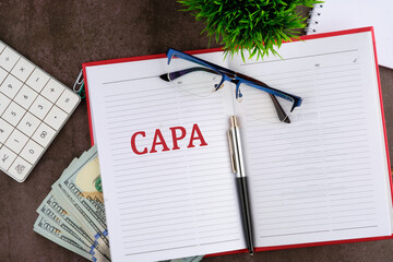 Concept image of Business Acronym CAPA Corrective and Preventive action written on the open page of the business notebook next to eyeglasses, a calculator, dollars, a plant and a pen.