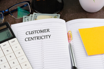 Customer Centricity word written in the manager's notebook in a composition with dollars, a calculator, a pen, stickers, glasses, paper clips