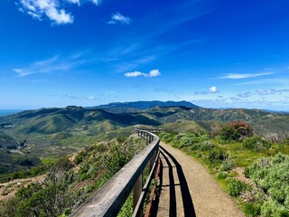 A narrow trail in the Marin headlands with a wooden rail in the foreground and blue sky above it