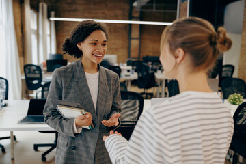 Two young diverse business women chatting during work break standing in modern office