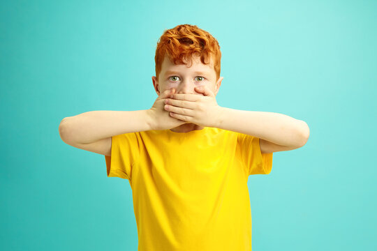 Little Red Headed Boy Covers Mouth With Hands, Looking to Camera, doesn't Want Say Anything, Wearing in Yellow T-shirt Standing Over Blue Isolated