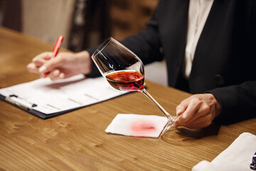Unrecognizable woman sommelier evaluates alcoholic drink, describes it with rating on paper. 