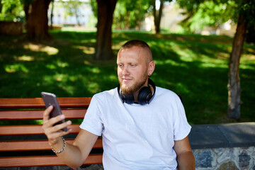 Fototapeta premium Man With Headphones Using Smartphone on Park Bench in Summer