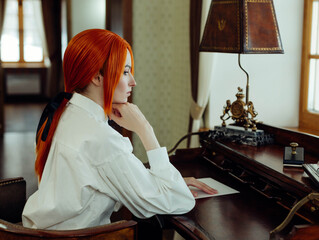 Red haired woman wearing a white blouse is sitting at a wooden desk, intently reading a letter, in...