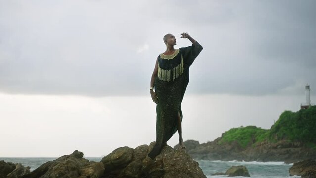 LGBTQIA black person in boutique dress, jewelry stands on rock on stunning ocean coastline skyline. Gender fluid ethnic divine model in luxury outfit fluttering in the wind against dramatic sky.