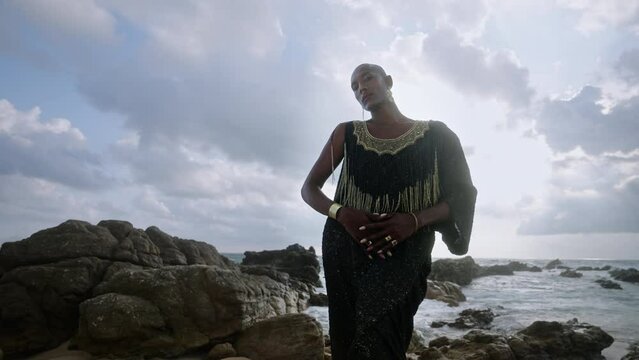 Trans sexual ethnic fashion model in long posh dress and accessories in elegant posture looks at camera slow motion. Epatage gay black man in luxury gown poses on stormy ocean beach, moody sky.