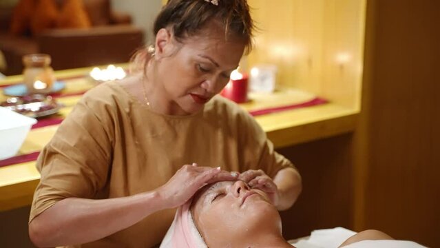 Senior multiracial masseuse cleances a young female client face with hands using facewash in spa. Adult worker of a wellness center performs a skincare procedure to a young woman on massage table