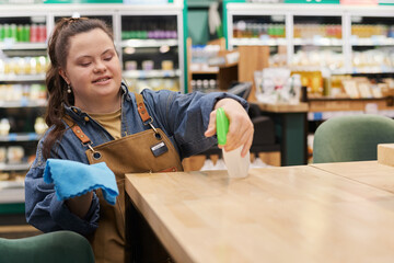 Waist up portrait of smiling young woman with disability working in supermarket and sanitizing...