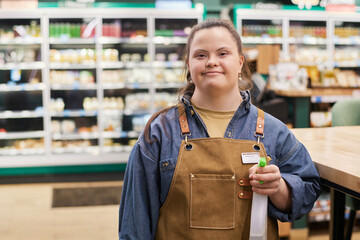 Waist up portrait of smiling young woman with Down syndrome working in supermarket and holding...