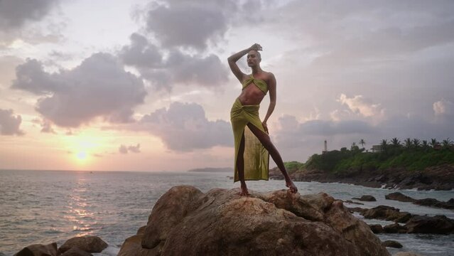 Godlike gender fluid person of color in open dress, jewelry stands on cliff peak at scenic ocean sunrise. Pride extravagant fashion model in luxury outfit posing like goddess in tropics. Olymp concept