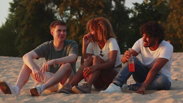 Three students telling funny situations in life when meeting the sunset. Best friends drinking alcohol on rivercoast. Summer time chill atmosphere. Humorous mates can't stop laughing