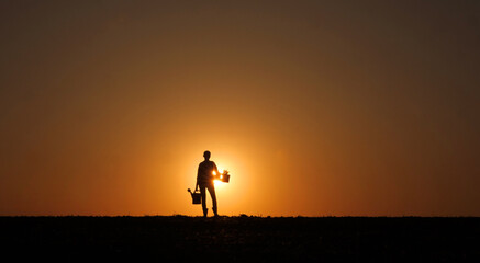 At sunset, a woman farmer stands in a field, her silhouette distinct as she holds a watering can and a seedling.