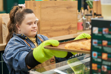 Portrait of young woman with Down syndrome working in bakery and pulling fresh croissants out of...