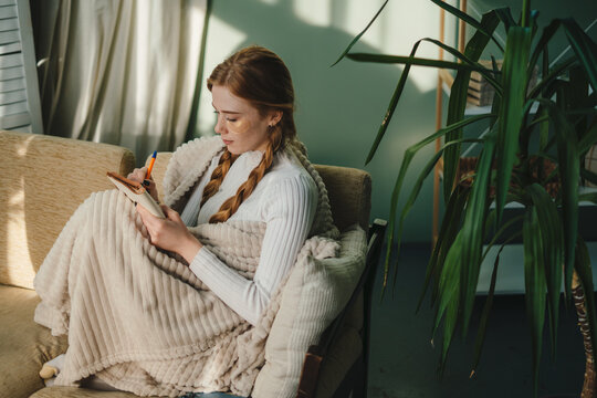 Ginger woman writing ideas in notebook, doing beauty face mask for skincare sitting in living room at home. Creative female looking thoughtful while planning