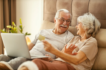 Cheerful senior couple in a bedroom holding a credit card and shopping online on a laptop.