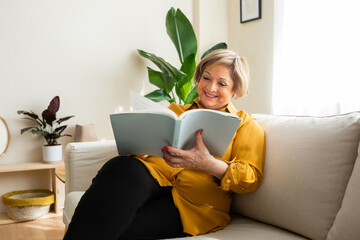 Smiling senior woman sitting in a living room reading a book. Senior woman spending his free time...