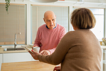 Elderly man holding a mug and chatting to his wife who is holding a tablet. Senior couple engaged...