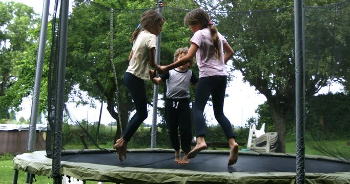 Super slow motion of carefree happy kids are having fun to jump together on elastic trampoline in garden outside his house on a sunny day. Childhood, family fun, freedom and happiness.