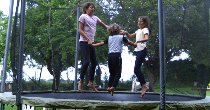 Super slow motion of carefree happy kids are having fun to jump together on elastic trampoline in garden outside his house on a sunny day. Childhood, family fun, freedom and happiness.