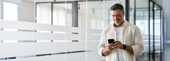 Banner with copy space. Smiling 40s mature Latin or Indian businessman holding smartphone in office. Middle aged manager man using cell phone mobile app. Digital technology application for business.