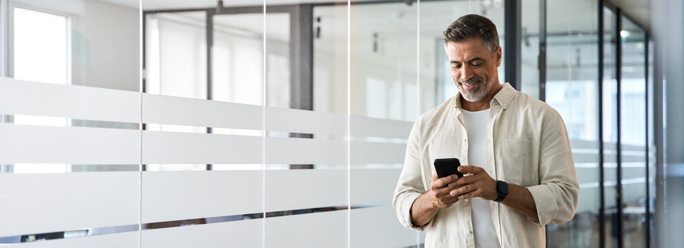 Banner with copy space. Smiling 40s mature Latin or Indian businessman holding smartphone in office. Middle aged manager man using cell phone mobile app. Digital technology application for business.