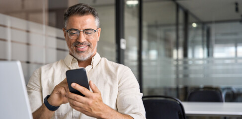 Banner with middle-age Hispanic business manager ceo using cell phone mobile app. Smiling Latin or Indian mature man businessman holding smartphone in office, working as trader on pc, with copy space