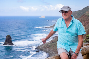 Handsome traveler senior man enjoying seaside holidays points with hand to famous Benijo beach in...
