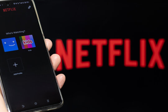 A hand showing the Netflix home screen, application logo blurred in the background. Streaming platform profile selection.