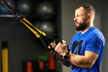 Serious strong man holding loops of trx equipment is training in gym. Concept of functional training