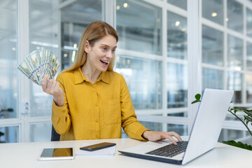Woman in a yellow blouse holding cash and using a laptop in a bright, modern office, expressing...
